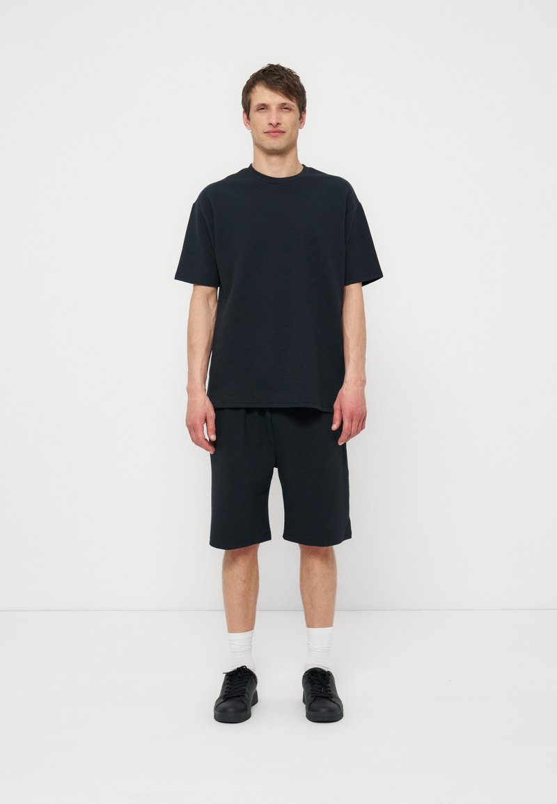 Νέος άνδρας που στέκεται σε λευκό τοίχο, φορώντας μαύρο oversized t-shirt, σορτς μήκους έως το γόνατο στο ίδιο χρώμα, λευκές κάλτσες και μαύρα αθλητικά παπούτσια.