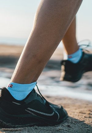 Scarpe sportive nere con un design elegante e dettagli grigi, abbinate a calzini alla caviglia azzurri chiari con un piccolo motivo rosso.