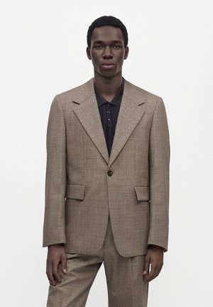 Homme portant un costume marron texturé avec un blazer à un bouton et un pantalon assorti, debout devant un fond blanc uni.