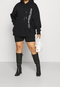 Felpa nera over-size con logo bianco, abbinata a pantaloni corti neri e stivali neri fino al ginocchio. Tiene una piccola borsa bianca.