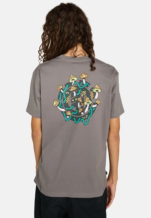 Personne avec de longs cheveux bouclés portant un T-shirt gris avec un graphique coloré de champignon et de vigne au dos, sur fond blanc.