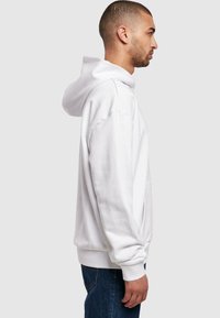 Weißer Hoodie mit Kängurutasche, gerippten Bündchen und verstellbarem Kordelzug an der Kapuze. Das Modell trägt dunkle Jeans und wird von der Seite gezeigt.