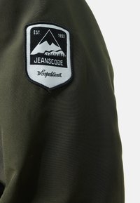 Giacca verde oliva in tessuto texturizzato. Presenta una toppa ricamata rotonda che mostra una montagna e alberi, con la dicitura "JEANSCODE" e "Expedition."