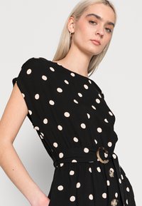 Schwarzes Kleid mit weißen Punkten, kurzen Ärmeln, tailliertem Schnitt und einem Gürtel mit runder Akzentuierung. Verfügt über eine Knopfleiste vorne und einen fließenden Stoff.