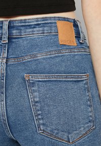 Jean en denim bleu à taille haute. Les caractéristiques incluent une étiquette en cuir portant la mention "PIECES", des coutures orange et deux poches arrière.
