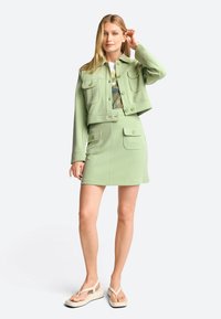Light green cropped jacket with front pockets, paired with a matching mini skirt. Features a smooth texture and button accents. Beige sandals.