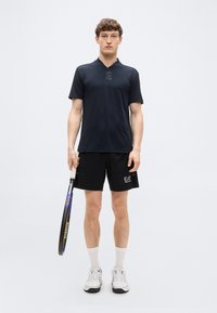 Ανδρικό αθλητικό σύνολο που αποτελείται από μαύρο πόλο με κοντά μανίκια, μαύρο σορτς και λευκά αθλητικά παπούτσια, κρατώντας ρακέτα τένις.