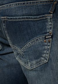 Jeans de denim con un lavado azul oscuro, que cuentan con un bolsillo trasero con detalles de costura y una pequeña etiqueta "GAS" cerca de los remaches metálicos.
