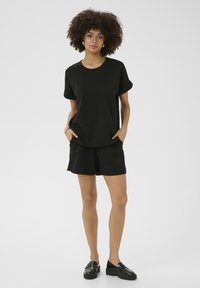 Juoda trumpa rankovė T-marškiniai su suktomis rankovėmis ir juodos šortai. Abu pagaminti iš lygios medžiagos. Stilius su juodais loafer'iais. Minimalistinis dizainas.