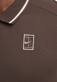 Hnedé polo tričko s textúrovaným povrchom, so širokým bielym golierom a rectangulárnym logom Nike v zlatej a bielej vyšitým na hrudi.