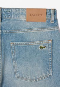 Šviesiai mėlynos džinsinės kelnės su odine etikete, pažymėta "LACOSTE", ir mažuoju žaliu krokodilu ant užpakalinio kišenės.