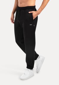 Mustat juoksuhousut pehmeästä materiaalista, joissa on joustava vyötärö, sivutaskut ja käärityt lahkeet. Yhdistettynä valkoisiin lenkkareihin.