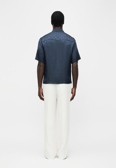 Άνδρας που στέκεται με την πλάτη στην κάμερα, φορόντας κοντομάνικο πουκάμισο σκούρου μπλε με σχέδια και φαρδιά λευκά παντελόνια, μπροστά σε απλό φόντο.