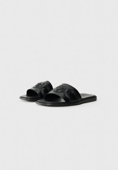 Svarta lädersandaler med slingback som har en framträdande cirkulär logotyp på remmen och texturerad detaljering längs sidorna. Platt sula.