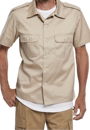 Hombre con camisa beige de manga corta con dos bolsillos delanteros con botones sobre una camiseta blanca y pantalones cortos cargo kaki.