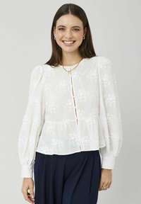Blouse blanche en tissu léger avec broderie florale, manches longues bouffantes et devant à boutons. Présente une taille évasée style peplum.