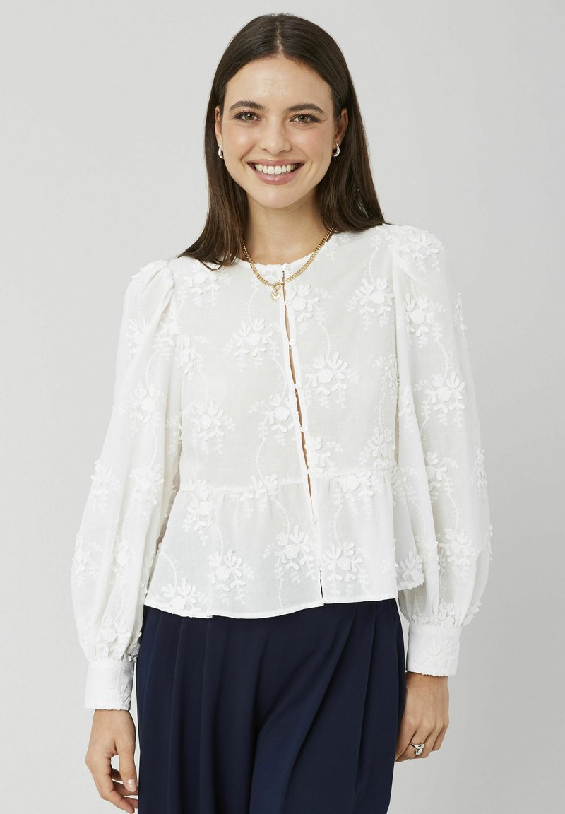 Blouse blanche en tissu léger avec broderie florale, manches longues bouffantes et devant à boutons. Présente une taille évasée style peplum.