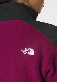 Fleecejacka i livlig lila med svarta axelpaneler. Har en broderad vit logotyp på vänster sida, texturerat tyg och hög krage.