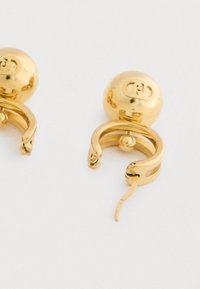 Egy pár fényes arany színű, gömb alakú fülbevaló, amelyeken gravírozott logók és zsanéros zárak találhatók, fehér háttér előtt.