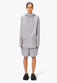Jasnoszary płaszcz przeciwdeszczowy z kapturem, zatrzaskami, bocznymi kieszeniami i sznurkami do regulacji. Połączony z jasnymi szortami i czarnymi butami.