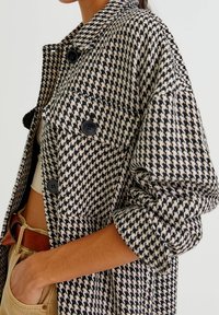 Zwart-wit oversized jas met pied-de-poule patroon, zwarte knopen, grote zakken en opgerolde mouwen, gemaakt van een gestructureerde stof.