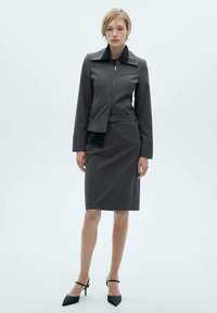 Maßgeschneiderter grauer Anzug mit einem taillierten Blazer mit scharfem Kragen und Reißverschluss, kombiniert mit einem figurbetonten Bleistiftrock; schicke schwarze Absatzschuhe.