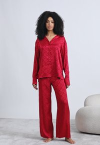 Czerwony satynowy zestaw piżamowy z długim rękawem i guziczkami, oraz szerokimi spodniami, oba elementy ozdobione geometrycznym wzorem i gładką fakturą.