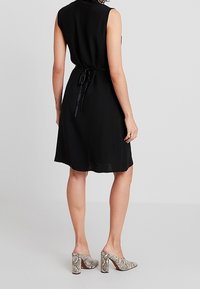 Schwarzes ärmelloses Kleid mit einem taillierten Design, fließendem Stoff und einer Bindeschleife am Rücken. Kombiniert mit grauen High-Heel-Mules im Schlangenprint.