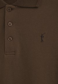Brun polo shirt i texturerat tyg, med en treknäppning och en broderad figur som svingar en golfklubba på vänster bröst.