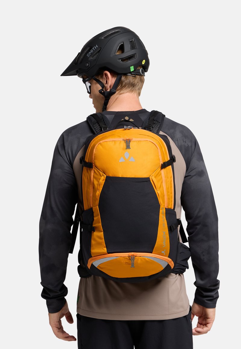 Sac à dos orange et noir avec plusieurs compartiments, texture en maille, fermetures éclair contrastées et accents réfléchissants, porté par une personne avec un casque.