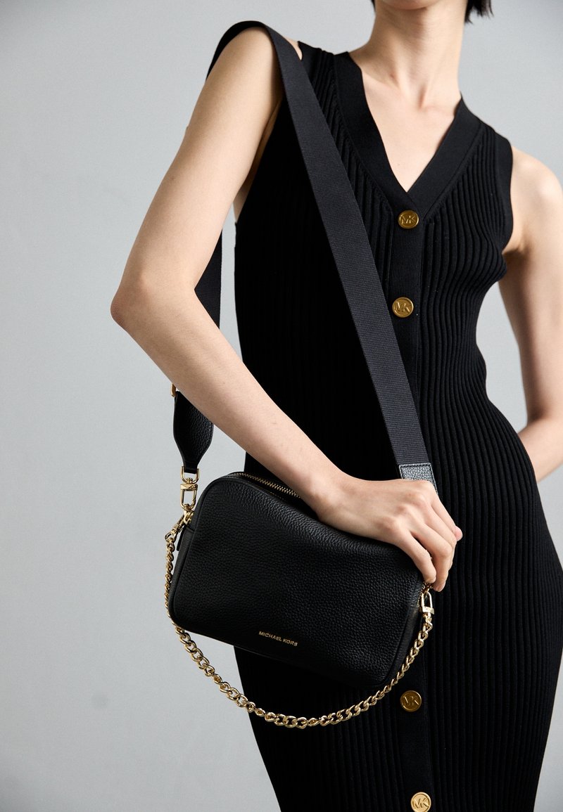 Černá kožená crossbody taška s texturovaným povrchem, zlatým řetízkem a zipovým uzávěrem. V kombinaci s černými žebrovanými šaty s zlatými knoflíky.