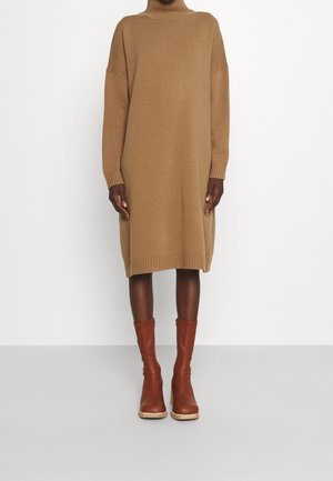 Person trägt ein langes, gestricktes Pulloverkleid in Kamelhaarfarbe mit Rollkragen und braunen Lederstiefeln bis zur Mitte der Wade vor einem schlichten weißen Hintergrund.