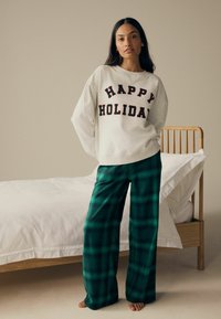Ljusgrå bomulls sweatshirt med "HAPPY HOLIDAY" tryckt i svart och rött; kombinerad med gröna rutig flanellbyxor. Träbädd i bakgrunden.