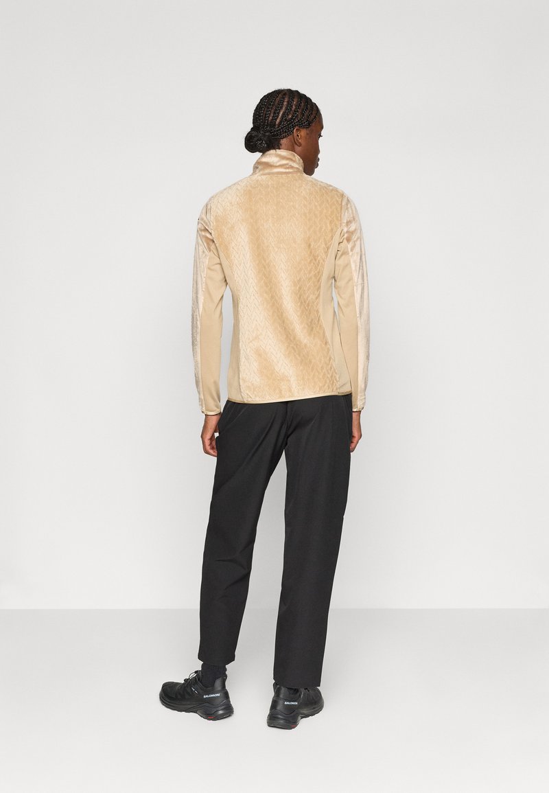 Beżowa, fakturowana bluzka z długim rękawem i wysokim kołnierzem, z delikatnym geometrycznym wzorem na plecach, sparowana z czarnymi spodniami i butami.