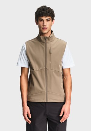 Mężczyzna ubrany w beżową kamizelkę bez rękawów zapinaną na zamek, na białą koszulę z krótkim rękawem i czarne spodnie, stojący na tle jednolitego jasnego tła.