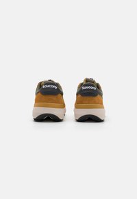 Saucony joggesko med brun semsket skinn, grønne detaljer og en kremfarget mellomsåle. Har en pull-tab og logo på hælen. Strukturert yttersåle.