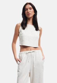Hvid og beige ternet crop top med firkantet halsudskæring og tynde stropper, parret med lyse beige, afslappede bukser med lommer.