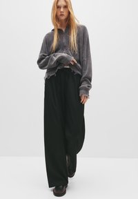 Sivý prúžkovaný oversized sveter s rozstrapkaným lemom v kombinácii s voľnými čiernymi nohavicami. Obuv má tmavú farbu a je v dizajne na obúvanie.