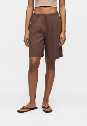 Donna che indossa pantaloncini marroni su misura fino al ginocchio, top corto beige e sandali infradito neri, in piedi davanti a uno sfondo uniforme.