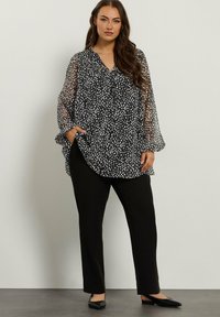 Donna che indossa una blusa trasparente a pois bianchi e neri con pantaloni neri e ballerine nere, in posa contro un muro chiaro uniforme.