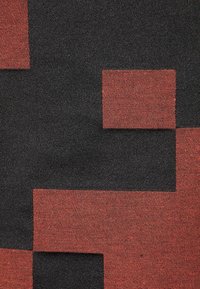 Textil s vzorem velkých geometrických červených a černých bloků s texturovaným, tkaným vzhledem. Pozadí je převážně tmavé.
