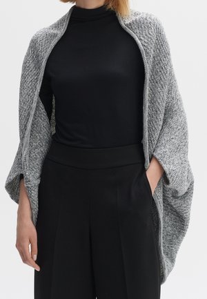 Vrouw in een zwarte coltrui en brede zwarte broek draagt een grijze gebreide sjaal over haar schouders, met één hand in de zak.