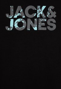 Jack & Jones Logo mit grauen Buchstaben und hellblauen Farbspritzern auf einem schwarzen Stoffhintergrund.