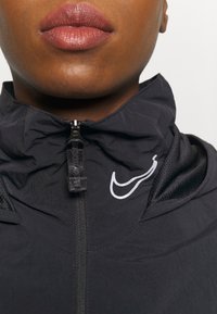 Black zip-up jacket with a high collar, featuring a silver Nike logo and textured fabric accents near the shoulders and neckline.