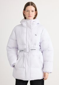Padnúci biely kabát s vysokým golierom, pásom s logom "Calvin Klein" a bočnými vreckami. Hladký povrch a prešívaný dizajn.