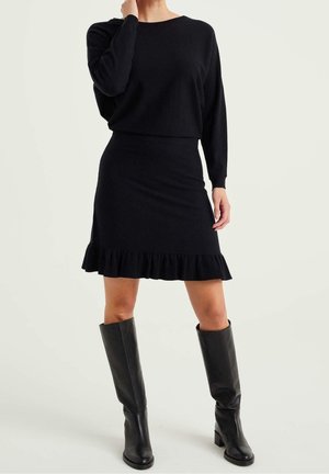 Femme portant un haut noir à manches longues et une jupe assortie à ourlet volanté, associée à des bottes en cuir noires montantes jusqu'aux genoux.