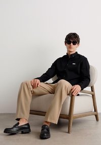 Zwarte lange mouw button-up shirt met een logo, beige broek en zwarte instappers. Zittend op een houten stoel op een neutrale vloer.