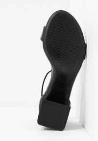 Semelle de sandale noire avec motif antidérapant texturé et petit talon bloc, vue de dessous sur fond blanc.
