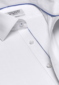 White dress shirt with herringbone texture, blue accent stitching along the collar, and rounded cuffs. Features pearl buttons and a label.
