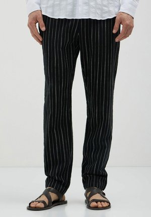 Pantalons rayés noirs en tissu doux, ornés de fines lignes verticales blanches. Associés à des sandales à lanières marron pour un look décontracté.